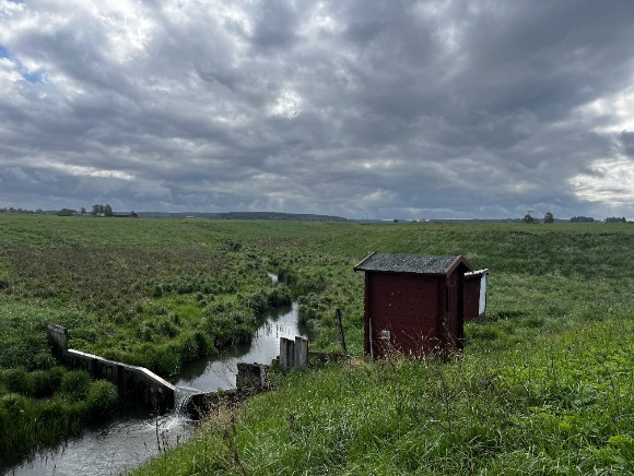 Vattendrag i ett jorbrukslandskap med ett litet rött hus bredvid.