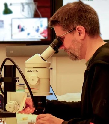 Man med brunt hår, skägg och svart tröja sitter och tittar i mikroskop