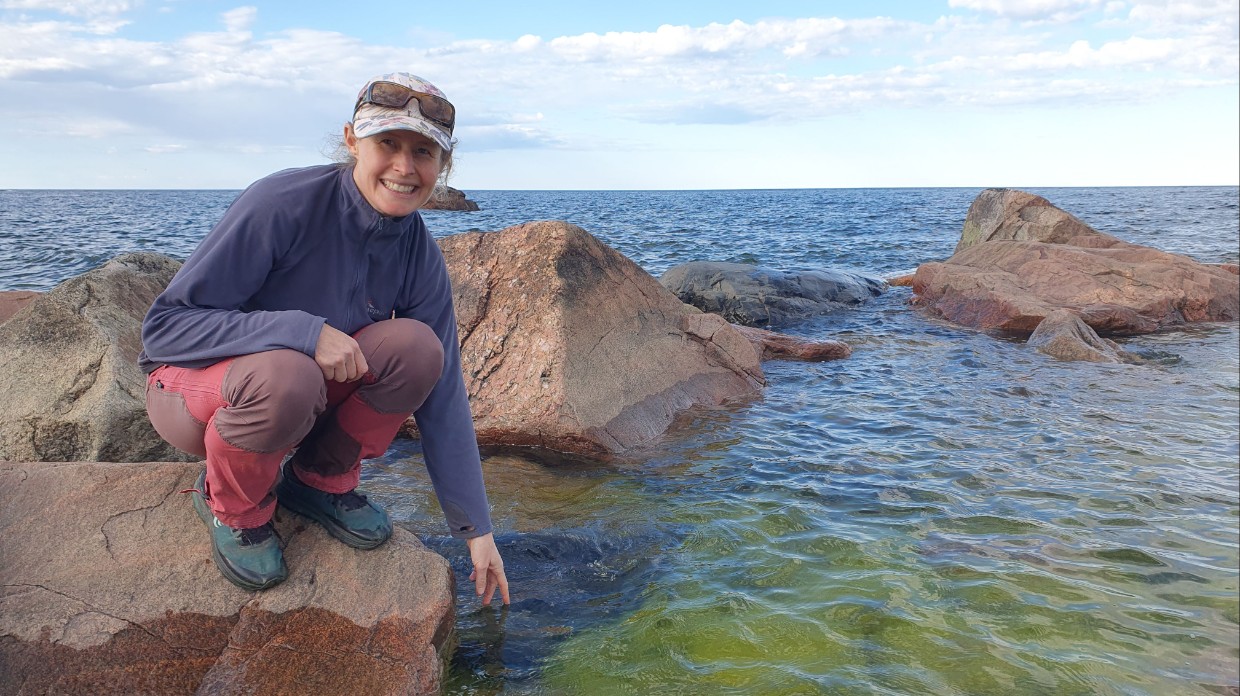 A woman sitting on a cliff by the sea reaches her hand down to touch the water. Photo. 