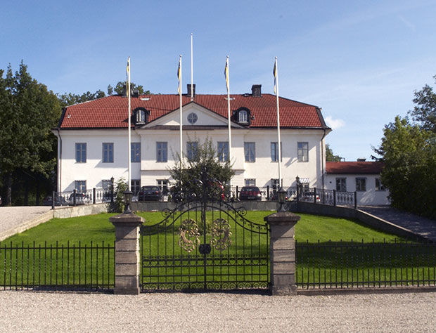 A white manor house in lovely summer weather, with four flagpoles and a large black iron gate in front.