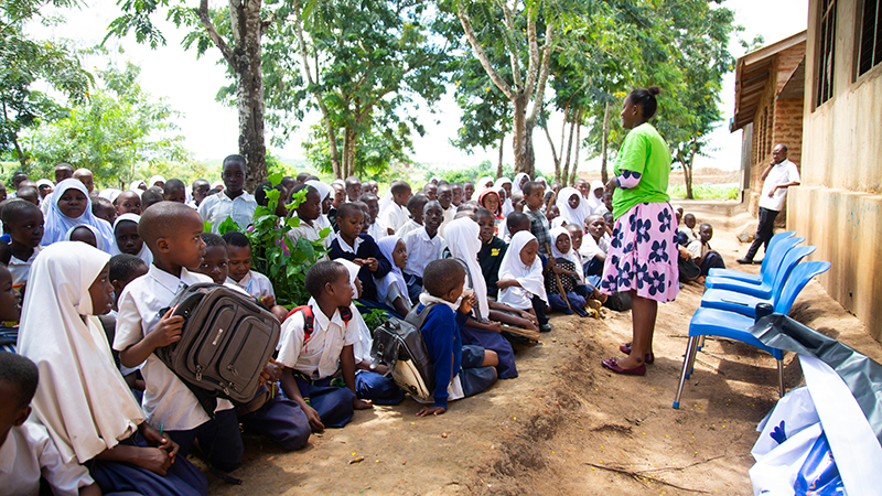An African country, where a woman stands in front of a group of people sitting on the ground and listening. Photo.