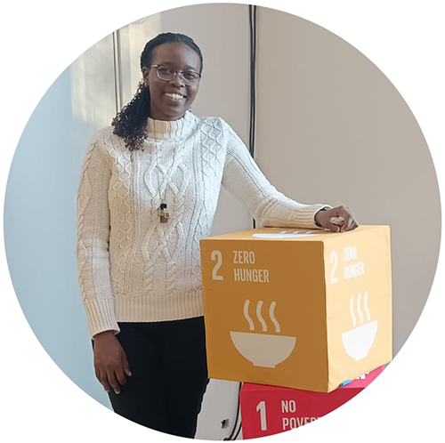 A smiling woman besides a yellow cube with the text “Zero hunger”. Photo. 
